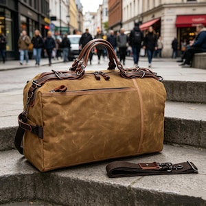 Puede incluir: Una bolsa de lona encerada marrón con detalles y asas de cuero. La bolsa tiene un compartimento principal con cremallera, un bolsillo lateral y una correa para el hombro desmontable. La bolsa está sobre escalones de piedra.