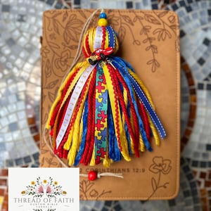 May include: A red, yellow, and blue tassel with a small red car charm and a white ribbon with the words "2 Corinthians 12:9" on it. The tassel is attached to a brown leather journal with the words "Thread of Faith Custom Bible Tassels" on a white label.