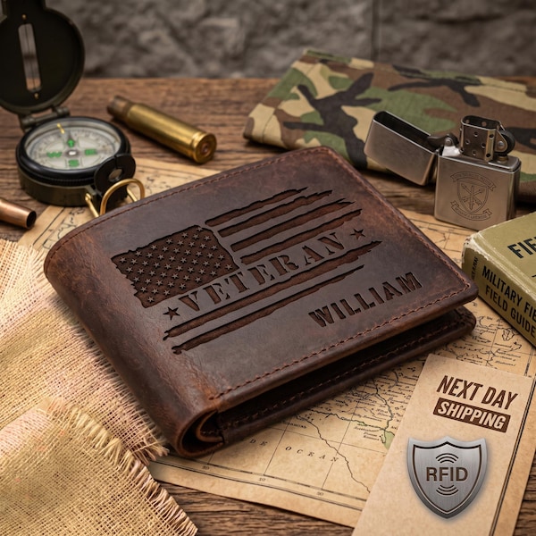 May include: A dark brown leather bifold wallet featuring a laser-engraved distressed American flag, the word VETERAN, and the name WILLIAM. The wallet is positioned on a map surface alongside a compass, a metal lighter, and a bullet casing.