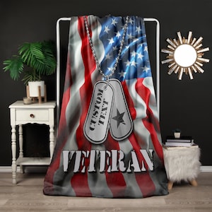 May include: A blanket featuring a waving American flag design with red and white stripes and blue stars. Two silver dog tags with the words "CUSTOM TEXT" and a star are suspended in front of the flag. The word "VETERAN" is printed at the bottom.