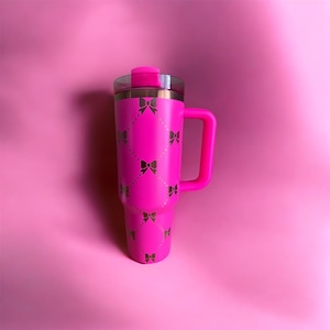 Pode incluir: Uma caneca de viagem rosa brilhante com uma alça e uma tampa transparente. A caneca é decorada com um padrão de laços dourados. O fundo é de cor rosa lisa.