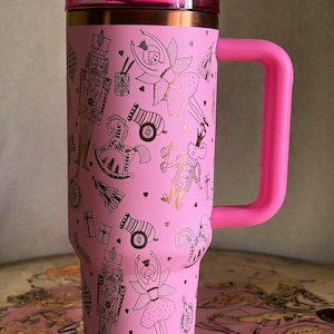 Könnte beinhalten: Rosa Reisebecher mit Henkel, mit einem gold-schwarzen Nussknacker-Motiv. Der Becher hat einen rosa Deckel und Henkel. Das Design umfasst Ballerinas, Zinnsoldaten und Kanonen.