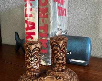 Könnte beinhalten: Ein braunes Salz- und Pfefferstreuer-Set aus Keramik mit zwei geschnitzten Tiki-Figuren auf einem Sockel, in dessen Mitte das Wort HAWAII eingeprägt ist. Der Sockel hat ein strukturiertes Blumenmuster und die Streuer zeigen detaillierte Gesichter.