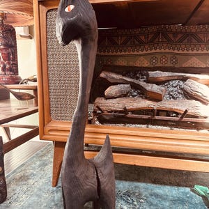 May include: A dark brown, hand-carved wooden sculpture of a stylized animal with a long neck and a pointed tail. The sculpture has a single eye with a red center. The wood grain is visible, adding texture to the piece. The sculpture is standing on a blue rug.