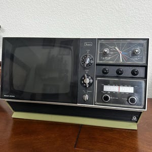 May include: A vintage Sears solid state television and radio combination unit with a black screen, analog clock, and AM/FM radio tuner. The device features silver knobs for VHF and UHF tuning, volume, contrast, brightness, and radio frequency selection.