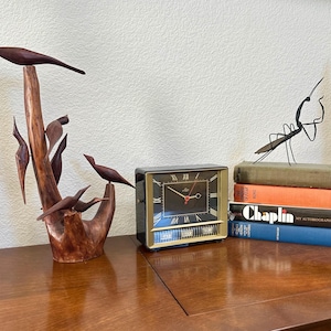 May include: A wooden sculpture of birds perched on branches, a vintage clock with a black face and gold trim, and a stack of books on a wooden surface. The clock displays the day and date.