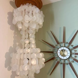 May include: A white capiz shell chandelier with a wooden top is suspended from the ceiling. The chandelier is composed of numerous round, translucent shells. A mid-century modern starburst clock with a gold and brown design is mounted on the wall.