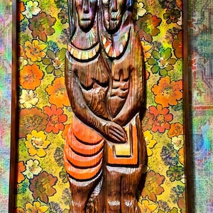 May include: A carved wooden relief sculpture of two figures, framed in a dark wood. The figures are depicted in a stylized, folk art style. The background is a floral pattern in shades of yellow, orange, and green.