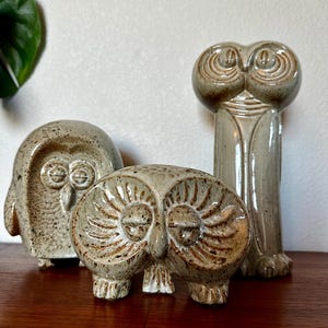 May include: Three ceramic owl figurines with a speckled, light beige glaze. One owl is tall and slender, while the other two are shorter and rounded. Each piece features carved details around the eyes and beak, showcasing a glossy, textured finish.