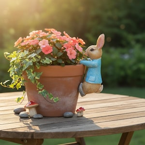 May include: A terracotta plant pot filled with pink flowers and trailing ivy. A decorative rabbit figure in a blue jacket clings to the pot's edge. Small mushroom and stone decorations are on the wooden table.