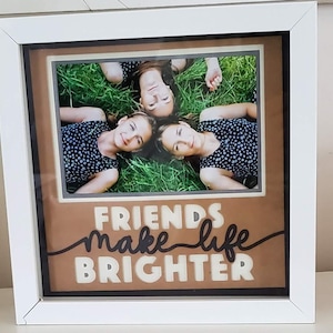 May include: White framed photo with three people lying in grass. The photo has a brown background with the words "FRIENDS make life BRIGHTER" in white and black lettering.