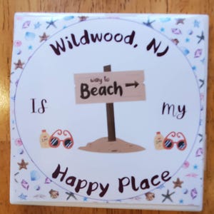 K&ouml;nnte beinhalten: Wei&szlig;er Keramikuntersetzer mit einem bunten Rand aus Muscheln und Sternen. Der Untersetzer zeigt den Text "Wildwood, NJ is my happy place" mit einem braunen Holzschild, das nach rechts zeigt und "way to Beach" sagt.
