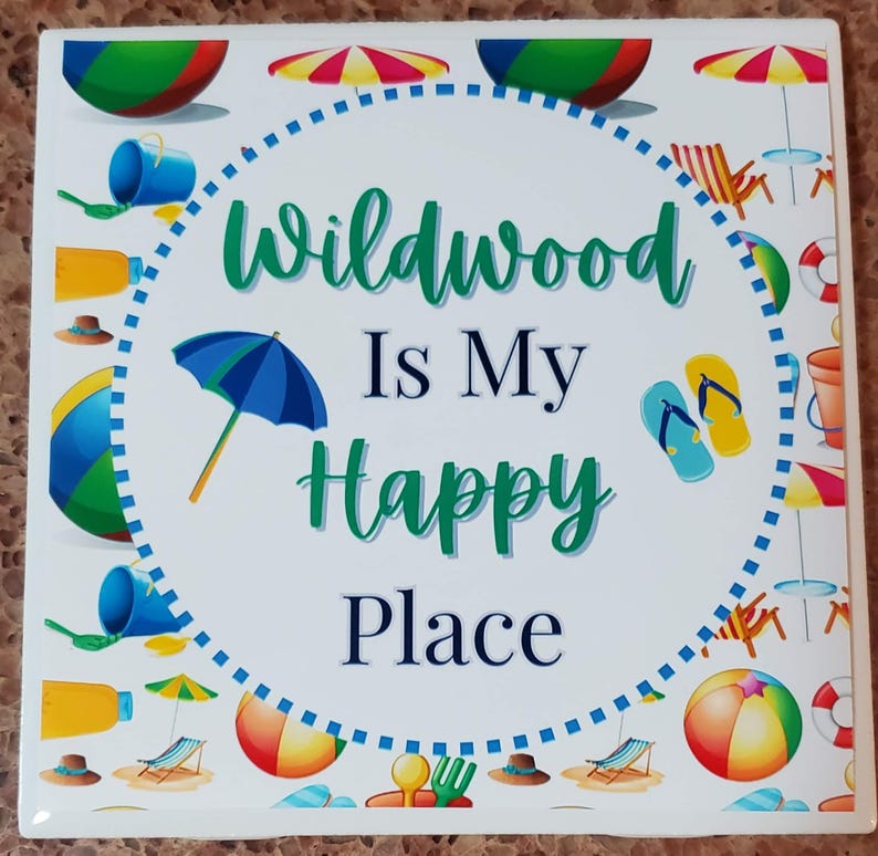 May include: White ceramic coaster with a blue and white dotted circle border. Inside the circle is the text "Wildwood Is My Happy Place" in green and blue lettering. The background is a white pattern of beach-themed items like beach balls, umbrellas, and flip-flops.