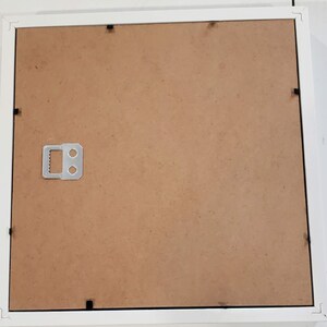 May include: A white picture frame with a brown backing. The frame has a simple, clean design with a square shape. A metal hanger is attached to the back for wall mounting. The frame is designed to hold a photograph or artwork.