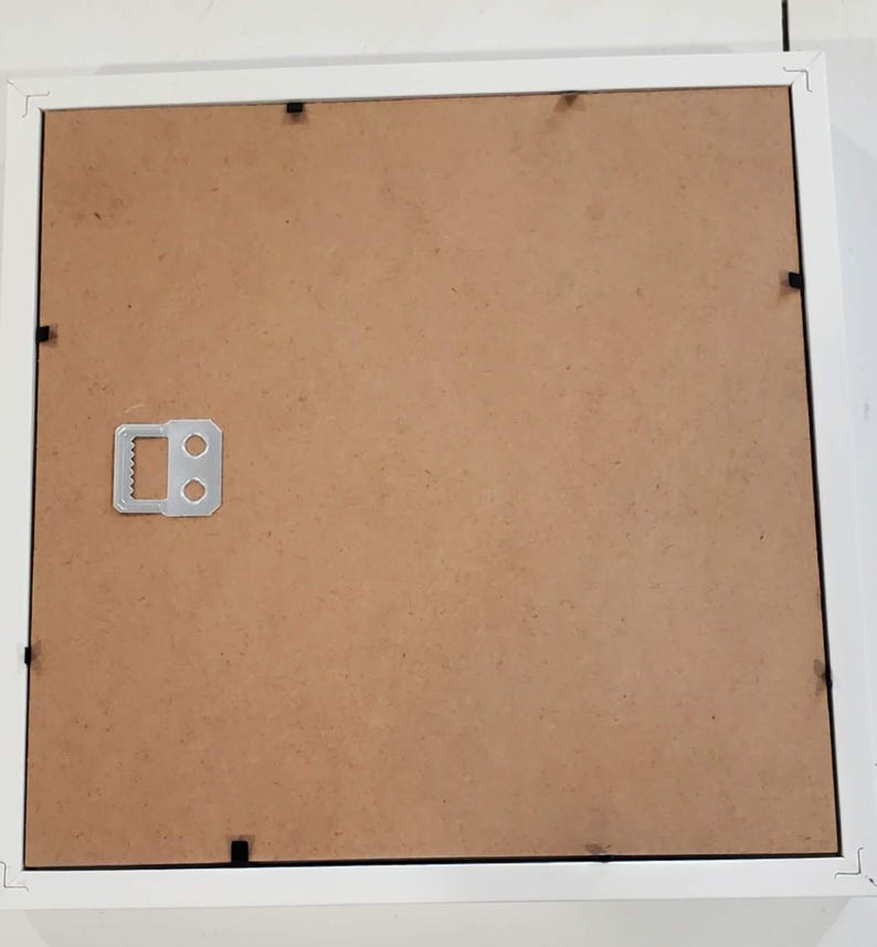 May include: A white picture frame with a brown backing. The frame has a simple, clean design with a square shape. A metal hanger is attached to the back for wall mounting. The frame is designed to hold a photograph or artwork.