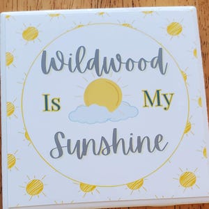 Pode incluir: Copo de cer&acirc;mica branco com um design de sol amarelo e nuvens. O texto "Wildwood is My Sunshine" est&aacute; escrito em letras cinzas.