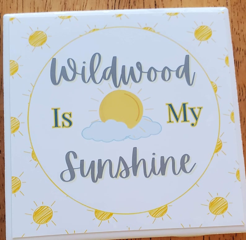 Pode incluir: Copo de cer&acirc;mica branco com um design de sol amarelo e nuvens. O texto "Wildwood is My Sunshine" est&aacute; escrito em letras cinzas.