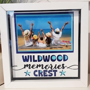 May include: White framed photo featuring a family on a beach with arms raised. The photo is surrounded by a blue border and the words "WILDWOOD memories CREST" in blue script and a star. The frame is square.