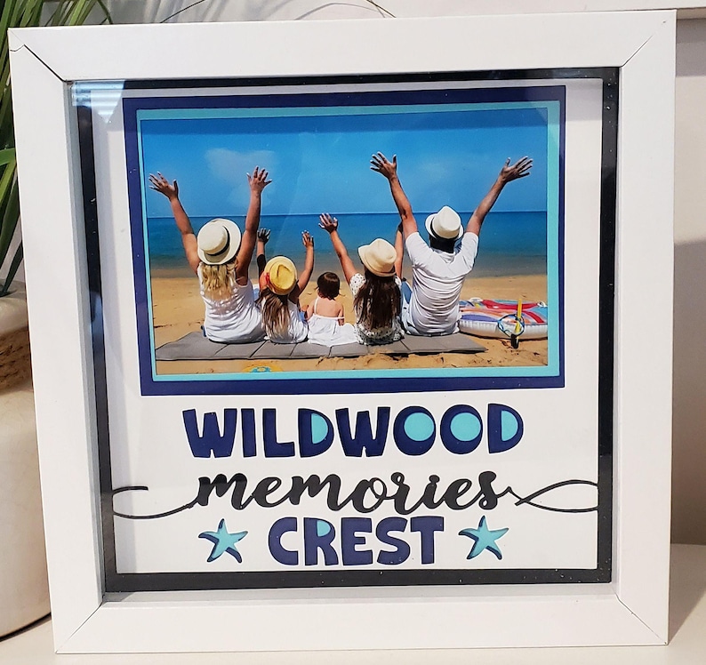 May include: White framed photo featuring a family on a beach with arms raised. The photo is surrounded by a blue border and the words "WILDWOOD memories CREST" in blue script and a star. The frame is square.