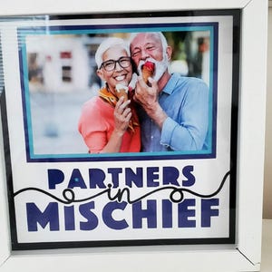 May include: Framed print featuring a smiling elderly couple enjoying ice cream cones. The image is framed in white with a blue and teal border. The text "PARTNERS in MISCHIEF" is displayed in blue and white.