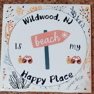 Puede incluir: Un azulejo de cer&aacute;mica blanco con un c&iacute;rculo negro en el centro. El c&iacute;rculo tiene el texto "Wildwood, NJ is my happy place" con una flecha rosa apuntando hacia la derecha que dice "beach". El azulejo est&aacute; decorado con un patr&oacute;n floral en negro, blanco, amarillo y naranja.