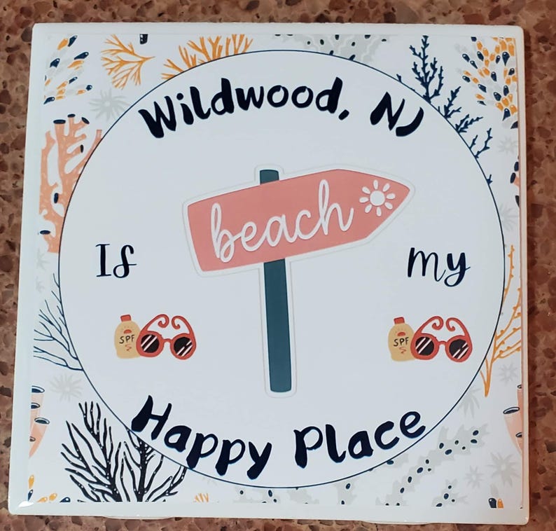 Puede incluir: Un azulejo de cer&aacute;mica blanco con un c&iacute;rculo negro en el centro. El c&iacute;rculo tiene el texto "Wildwood, NJ is my happy place" con una flecha rosa apuntando hacia la derecha que dice "beach". El azulejo est&aacute; decorado con un patr&oacute;n floral en negro, blanco, amarillo y naranja.