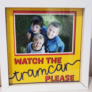 以下が含まれることがあります： 3人の笑顔の子供たちの写真が入った白いフレーム。写真は、赤と青の文字で「WATCH THE tramcar PLEASE」と書かれた黄色の背景に設定されています。フレームは白です。