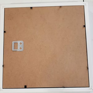 May include: A white picture frame with a brown backing. The frame has a square shape and a simple design. A metal hanger is attached to the back for wall mounting. The frame is designed to hold a photograph or artwork.