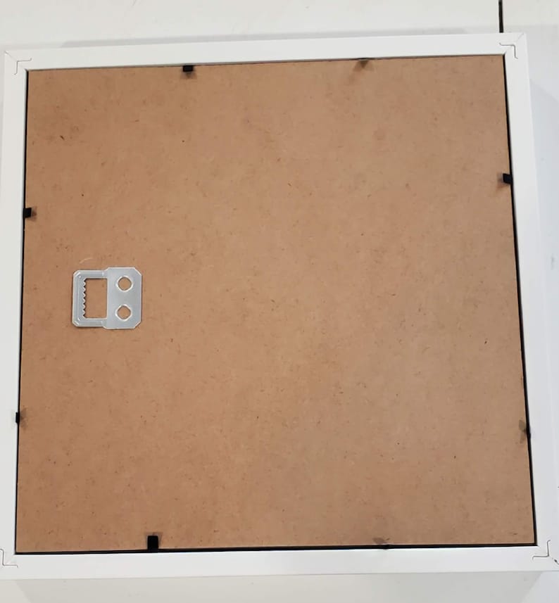 May include: A white picture frame with a brown backing. The frame has a square shape and a simple design. A metal hanger is attached to the back for wall mounting. The frame is designed to hold a photograph or artwork.