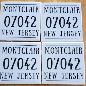 May include: Four white square tiles with black text. Each tile reads "MONTCLAIR" above "07042" and "NEW JERSEY" below. The tiles are arranged in a 2x2 grid on a wooden surface.