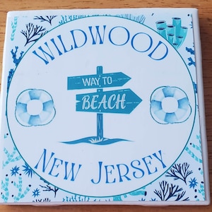 May include: Square ceramic coaster with a beach theme. The coaster features the words "Wildwood New Jersey" in blue, surrounding a central design with a signpost pointing "Way to Beach." Two blue and white life preservers and aquatic plant illustrations complete the design.