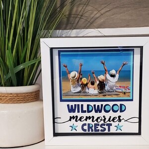 May include: A white framed photo with a beach scene featuring a family with their arms raised. The photo frame has the words "WILDWOOD memories CREST" in blue and white. A potted plant with green leaves is to the left of the frame.