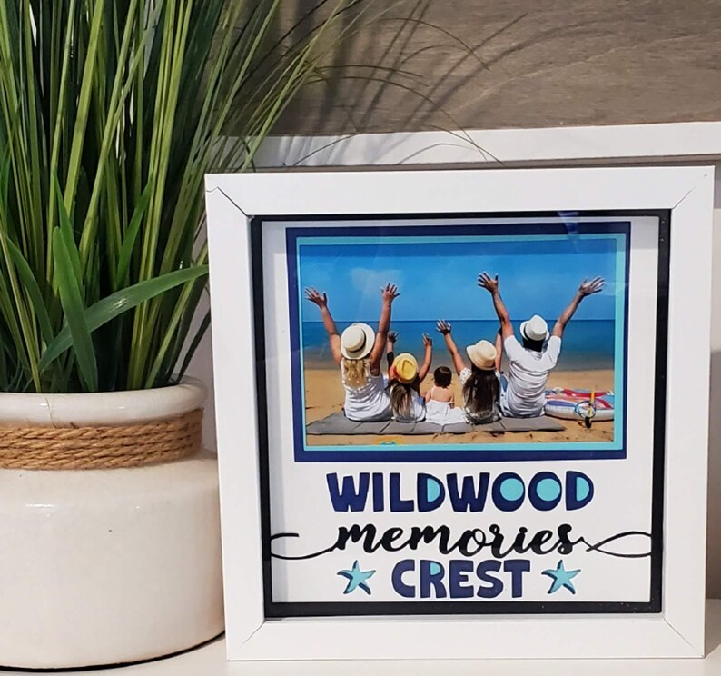 May include: A white framed photo with a beach scene featuring a family with their arms raised. The photo frame has the words "WILDWOOD memories CREST" in blue and white. A potted plant with green leaves is to the left of the frame.