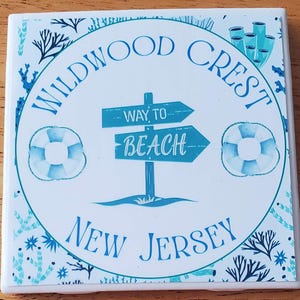 May include: Square ceramic coaster with a beach theme. The coaster features the words "Wildwood Crest New Jersey" in blue, along with a sign that says "Way to Beach" and two life preservers. The design includes blue and teal accents.