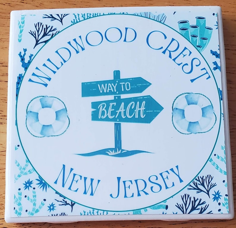 May include: Square ceramic coaster with a beach theme. The coaster features the words "Wildwood Crest New Jersey" in blue, along with a sign that says "Way to Beach" and two life preservers. The design includes blue and teal accents.