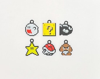 Op de afbeelding: Zes pixelachtige bedels met personages uit een videogame. De bedels bevatten een witte geest met een rode mond, een gele vraagtekenblok, een zwarte en grijze Bullet Bill, een gele ster, een rood-witte Koopa-schaal en een bruine Goomba.