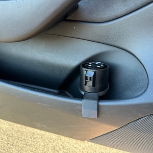 May include: A black plastic adapter with multiple ports is mounted to the inside of a car door. The adapter is attached to a black plastic bracket.