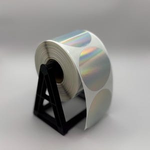 以下が含まれることがあります： 黒いプラスチック製のディスペンサーに、虹色の銀色の丸いステッカーが巻かれたロール。ステッカーは円形に配置され、ホログラフィックな仕上げになっています。