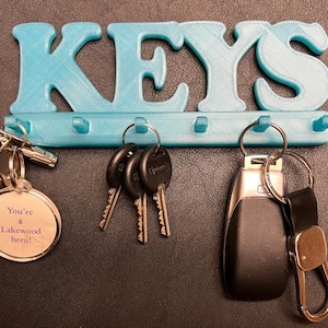 May include: A turquoise 3D-printed key holder with the word "KEYS" across the top. Below are five hooks holding keys and keychains. One keychain reads "You're a Lakewood hero!"