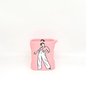 Op de afbeelding: Een lichtroze siliconen beschermhoes voor draadloze oordopjes met een grafische illustratie van een persoon met donker haar in een roze overhemd en een witte broek met hoge taille en knopen. De hoes heeft een kleine lus aan de zijkant.