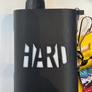Op de afbeelding: Een zwarte rechthoekige beschermhoes met het woord HARD uitgesneden in een vetgedrukt lettertype, waardoor een witte binnenkant zichtbaar is. De hoes heeft een zwarte bevestigingslus in de rechterbovenhoek, met een metalen clip en een gele band.