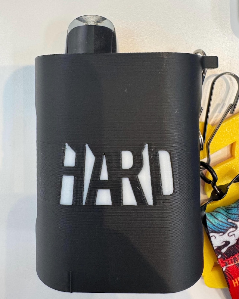 Op de afbeelding: Een zwarte rechthoekige beschermhoes met het woord HARD uitgesneden in een vetgedrukt lettertype, waardoor een witte binnenkant zichtbaar is. De hoes heeft een zwarte bevestigingslus in de rechterbovenhoek, met een metalen clip en een gele band.