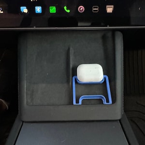 Pode incluir: Um suporte de plástico azul para uma caixa de carregamento sem fio branca, que está em um console de carro preto.