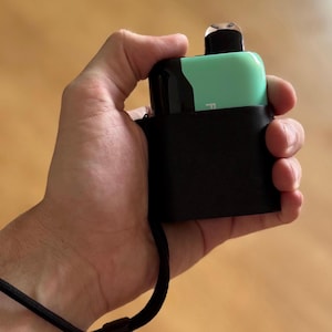 Op de afbeelding: Een rechthoekig elektronisch apparaat met een mintgroene en zwarte behuizing, voorzien van een transparant mondstuk aan de bovenzijde. Het apparaat zit in een zwarte hoes met een bevestigd zwart koord voor draagbaar gebruik.