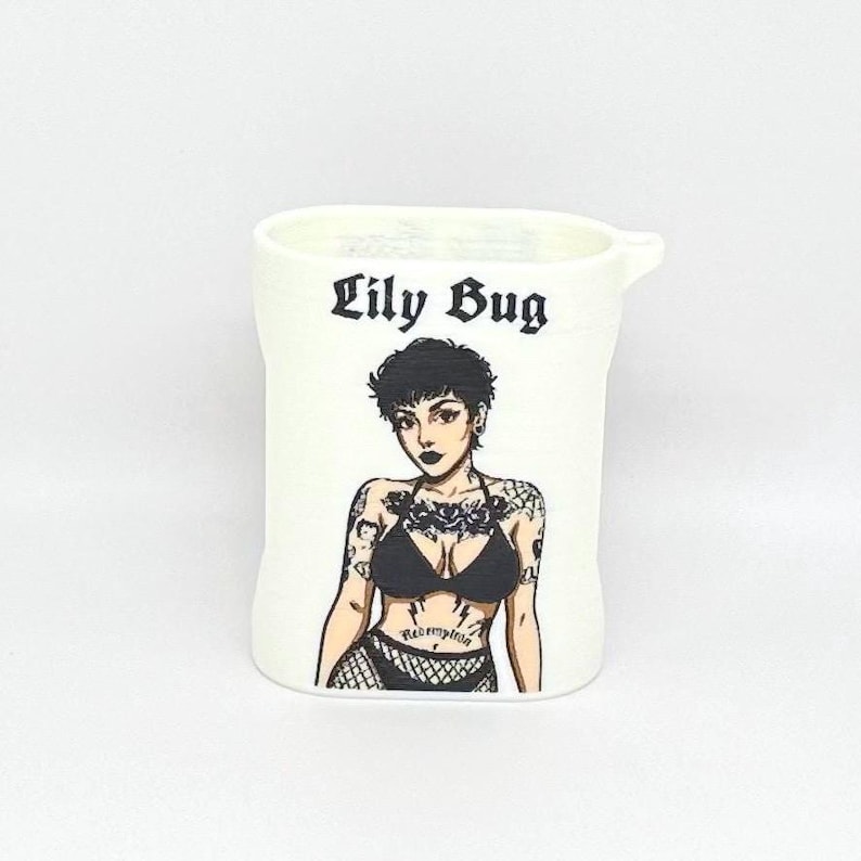 Op de afbeelding: Een witte cilindrische houder met de zwarte tekst Lily Bug boven een grafische illustratie van een persoon met kort donker haar, tatoeages op de armen en borst, gekleed in een zwart bikinitopje en visnet ondergoed met het woord Redemption.
