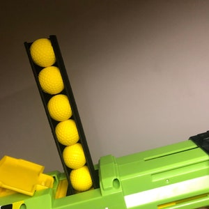 May include: A green and black toy gun with a yellow magazine loaded with yellow foam darts. The magazine is raised and the darts are visible.