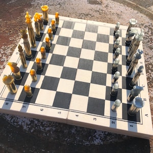 May include: A unique chess set with a wooden board featuring black and white squares. The chess pieces are designed to resemble bullets, with gold and silver tones. The board is marked with numbers and letters for gameplay.