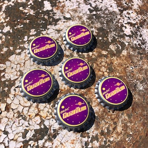 May include: Six metal bottle caps with a purple and yellow design. The caps feature the text "Quantum" and a planet graphic. The caps are arranged on a weathered, rusty surface.