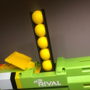 Può includere: Una pistola giocattolo Nerf Rival verde e nera con un caricatore giallo carico di palle di schiuma gialle. Il caricatore è etichettato "NERF RIVAL".