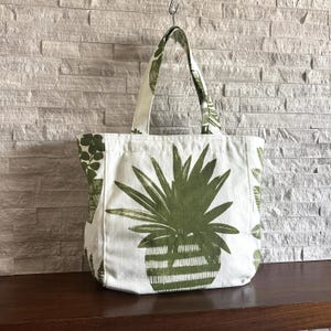 May include: White tote bag with a large green plant design. The bag features a large green plant in a striped pot on the front, with smaller plant patterns on the sides and straps. The bag is hanging from a metal hook.